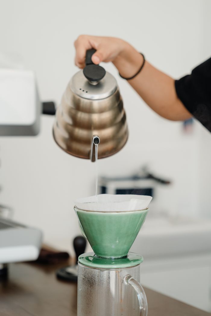 Crop unrecognizable female barista brewing coffee alternatively by pouring hot water from kettle into pourover in coffee shop environment