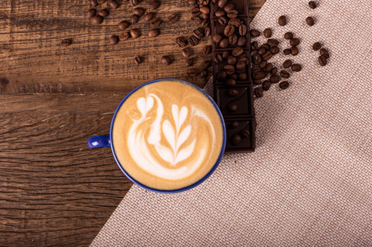 Overhead view of a latte with swan latte art, surrounded by coffee beans and chocolate on rustic wood.